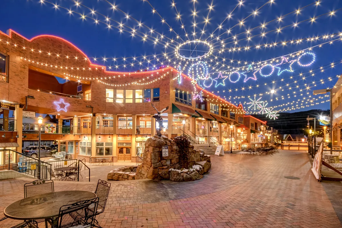 A vibrant downtown streetscape illuminated by decorative string lights, with inviting brick buildings, outdoor seating areas, and a prominent fountain or rock feature. People can be seen dining, socializing, and enjoying the lively atmosphere of the area.