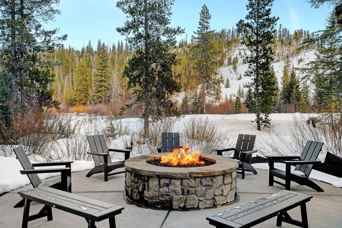 An inviting outdoor fire pit surrounded by comfortable seating, set amidst a snow-blanketed landscape with tall pine trees. The group of people gathered around the fire evokes camaraderie and après-adventure relaxation.