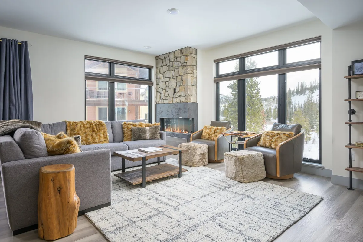 The living room features a welcoming stone fireplace, large windows framing serene forest views, and comfortable seating arranged for relaxation and socializing. A textured rug and cozy decor create an inviting mountain-chic atmosphere, perfect for gathering after outdoor adventures.