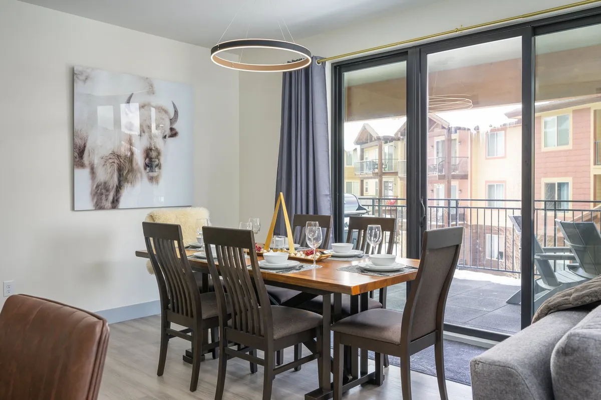 A custom wood dining table anchors the dining area, complemented by distinctive bison artwork and large glass doors that bring in natural light. The setting is ideal for shared meals, laughter, and connection.