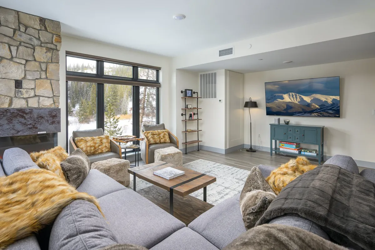 The image displays a cozy living area with a striking stone fireplace as the centerpiece, surrounded by plush, comfortable seating. Expansive windows flood the space with natural light and reveal scenic mountain views, while a wooden coffee table and a wall-mounted TV complete the inviting atmosphere. The mood is warm and welcoming, perfect for relaxation and gathering with friends or family.