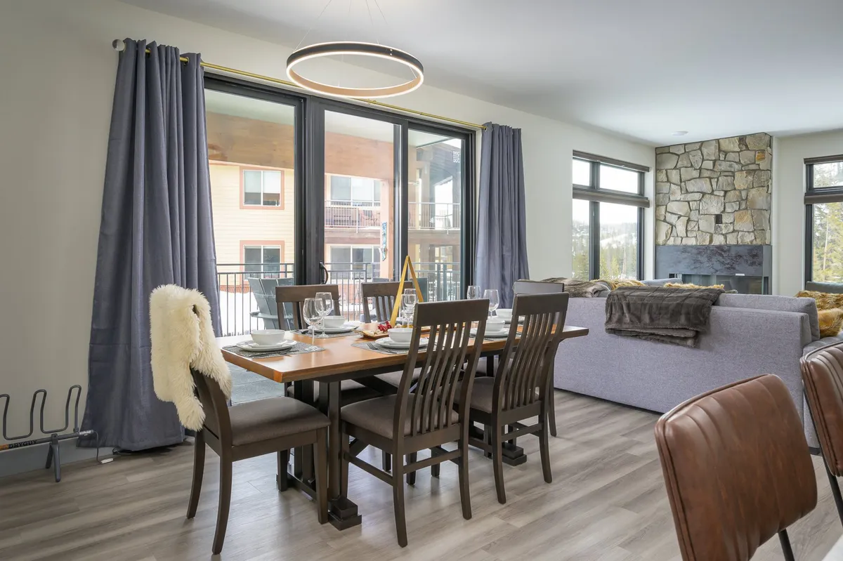 A bright, welcoming dining area with a large table set for dinner, expansive windows letting in ample natural light, and a cozy stone fireplace—suggestive of both relaxation and scenic views.