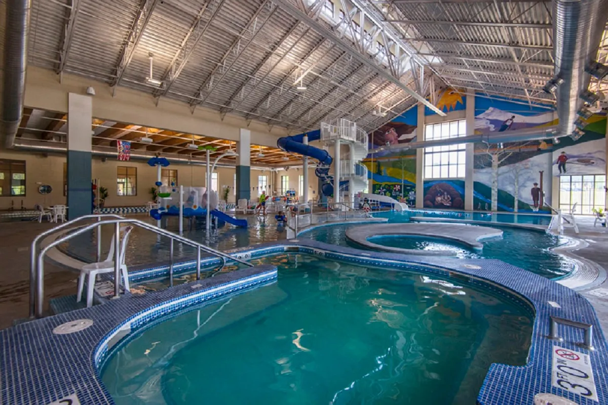 This image captures a lively indoor pool area at the Grand Park Rec Center, featuring a colorful water slide, children playing in the water, lounging chairs, and a vibrant mural on the wall. The scene is filled with energy and activity, representing family fun and wellness amenities available to guests.