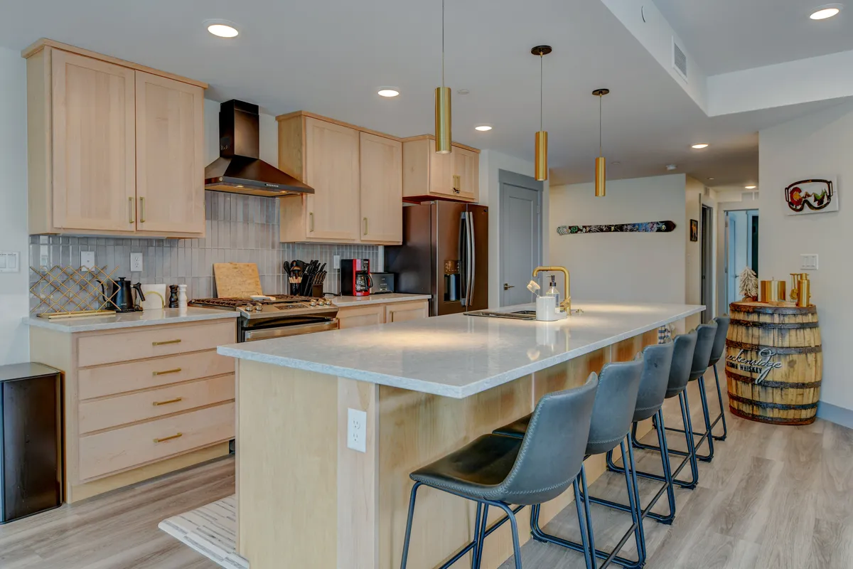 A modern gourmet kitchen with a spacious island, sleek pendant lights, state-of-the-art appliances, and rich wood cabinetry. The setup encourages both cooking and conversation, inviting group meal prep or festive cocktail sessions.