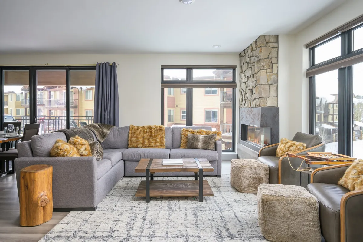 A spacious living area with a large sectional sofa, stone fireplace, wooden coffee table, plush ottomans, and textured throw pillows. The scene suggests a warm, inviting environment ideal for relaxing, socializing, and playing games together.