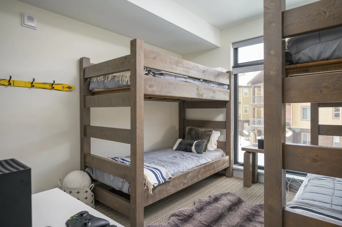 A playful bunk room featuring sturdy bunk beds, a textured rug, and a window filling the area with natural light. The space is inviting for kids and teens, with opportunities for sleeping, playing video games, or reading.