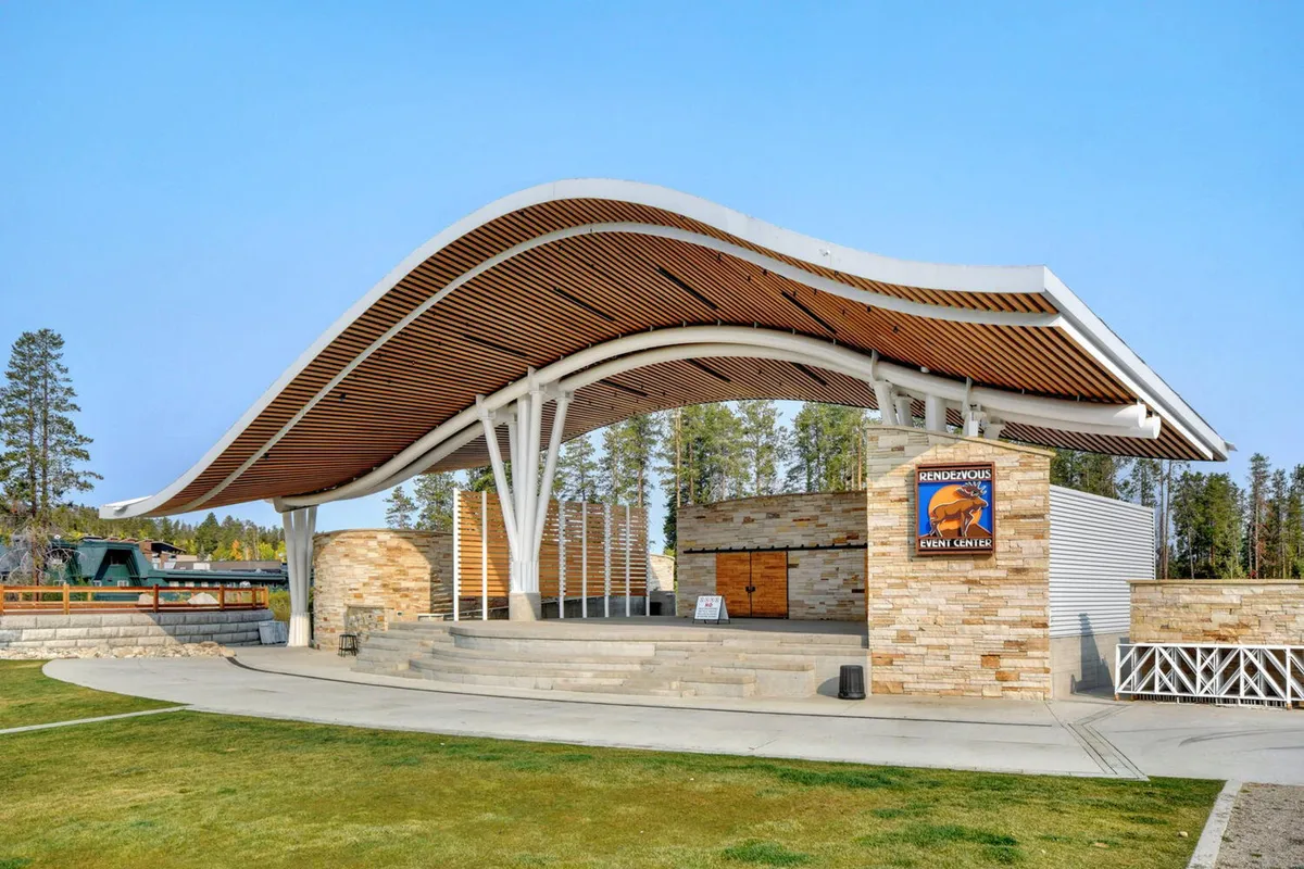 The Hideaway Park Amphitheater with its distinctive curved roof structure and stone façade, set up for an outdoor concert or community event. The stage area is visible, and the setting exudes a celebratory mood with the potential for live performances and gatherings.