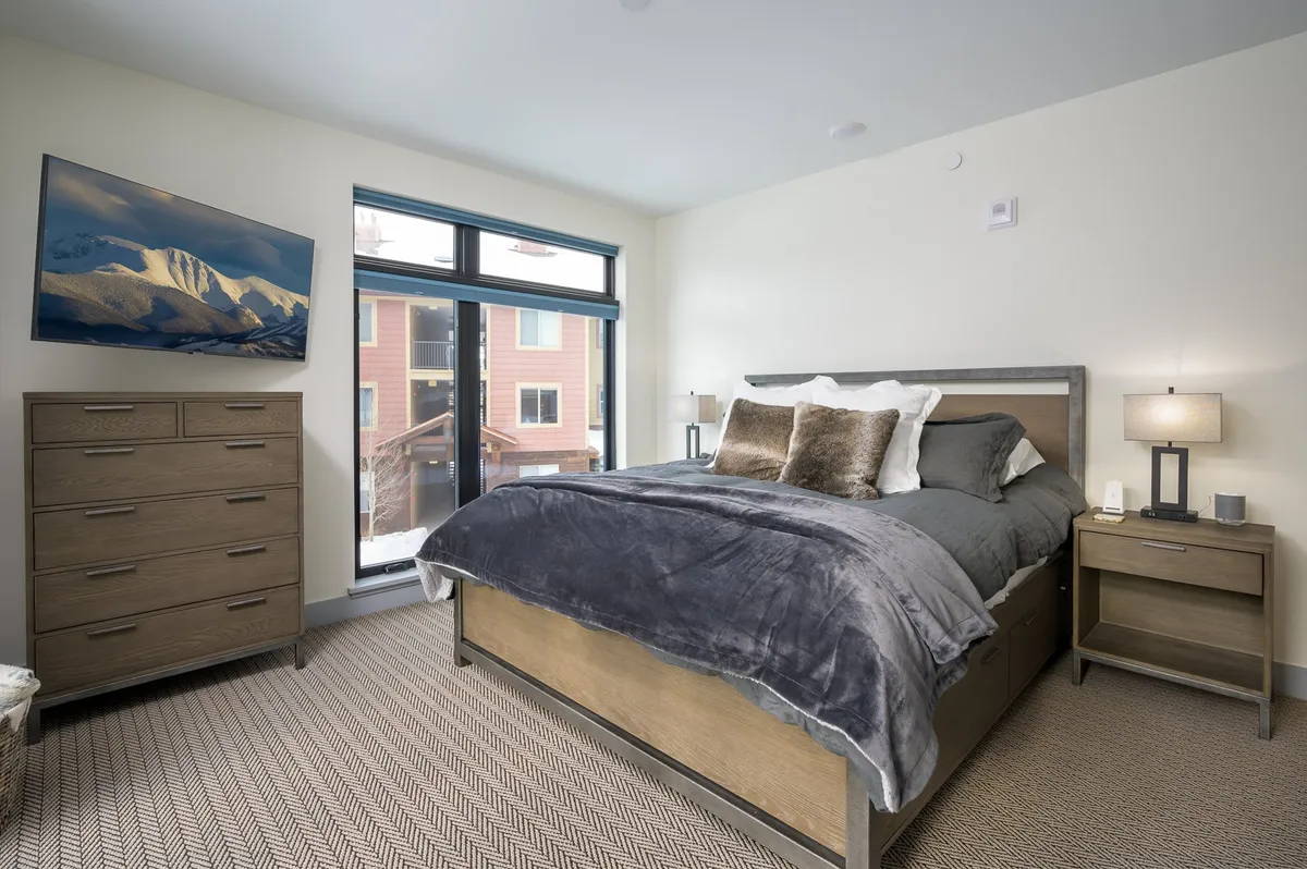 A serene bedroom featuring a neatly made bed with plush linens, a large window letting in natural light, a dresser, and thoughtfully chosen artwork on the walls. The space is uncluttered and exudes a calm, restful vibe, emphasizing cleanliness and comfort.