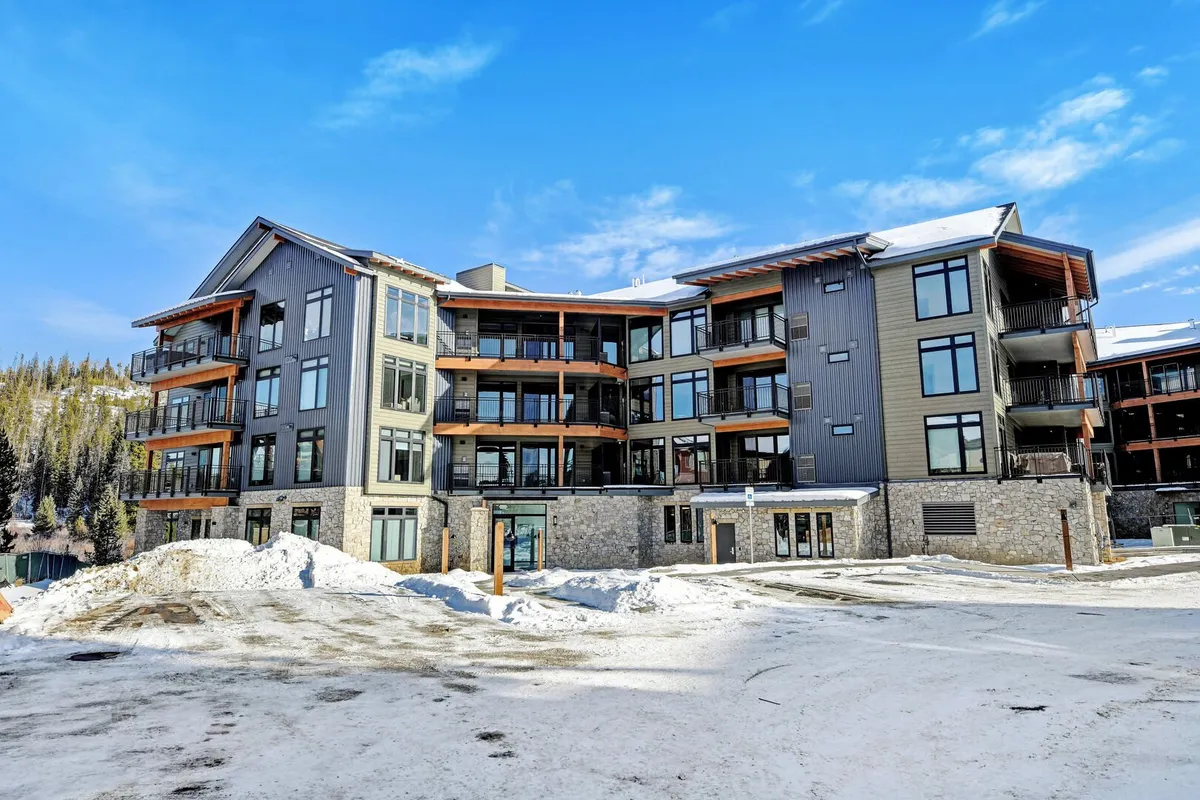 A view of the modern condo building set amid a snow-covered landscape, with large windows, wooden architectural accents, and balconies overlooking the mountains. The scene is welcoming, with hints of outdoor activities like skiing and relaxing on balconies.