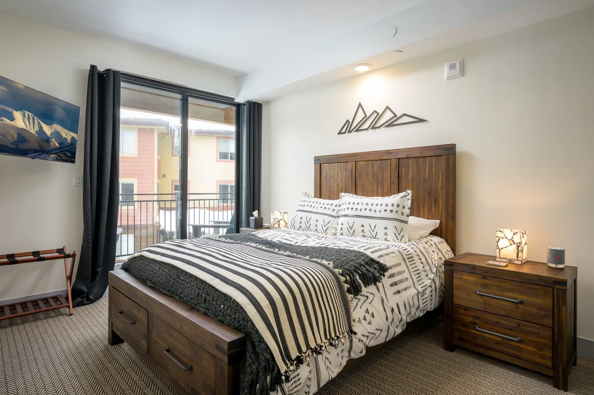 A cozy queen bedroom with patterned bedding, wooden nightstands, a TV mounted on the wall, and large windows with scenic views. Decorative mountain art adds a touch of character.