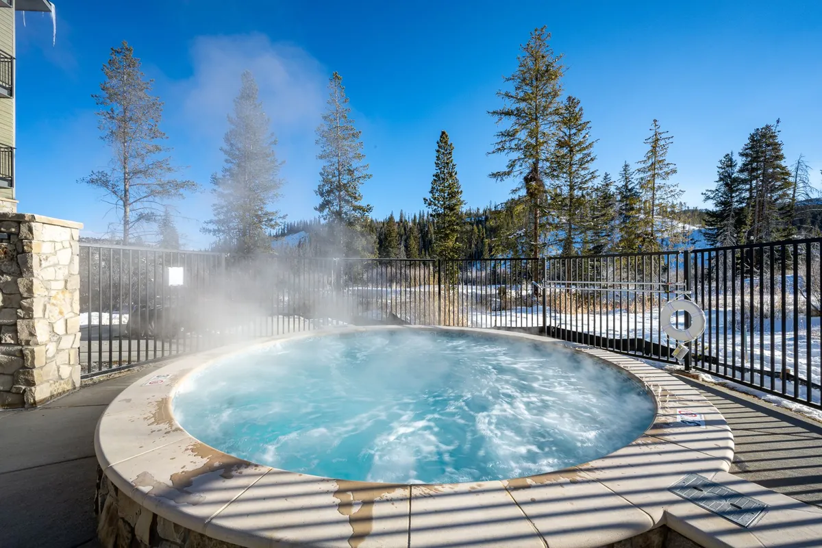 An inviting outdoor communal hot tub surrounded by trees with a backdrop of mountains. The hot tub is steaming, and the area is tranquil, evoking relaxation after a day of adventure.